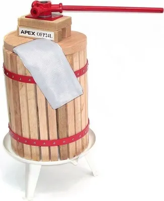 A wooden fruit press with a red handle and red bands, standing on a white base with legs. A white mesh bag is placed on top.