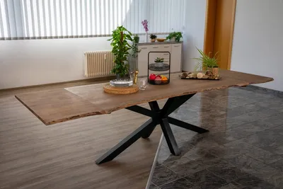 A dining table with a live edge wooden top and a black spider leg base, set in a dining room with blinds and cabinets in the background.