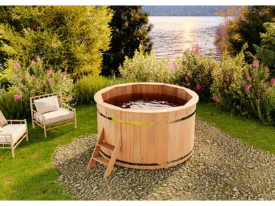 A wooden hot tub is set up in a garden next to a lake, with chairs and plants around it.
