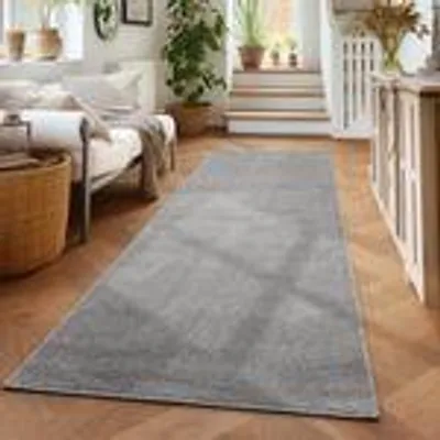 A rug is placed on a wooden floor in a living room setting, with a couch and plants visible.