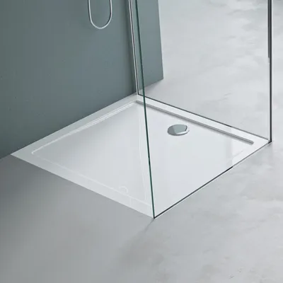 A white shower tray with a chrome drain is shown in a modern bathroom setting with gray walls and a concrete floor.