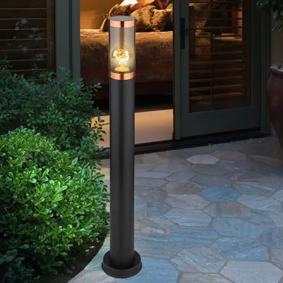 A black bollard light with a copper accent and a clear glass shade containing a light bulb stands on a stone patio next to lush green bushes. The background shows a doorway leading into a dimly lit room.