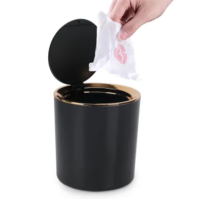 A hand is shown dropping a tissue with a lipstick kiss mark into a small black trash can with a gold rim and an open lid.