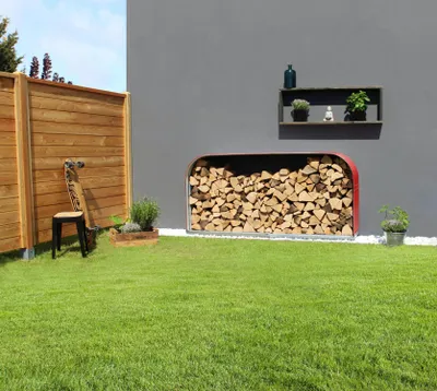 A modern outdoor space with a wooden fence, a lawn, and a grey wall featuring a firewood storage unit and a shelf with decorative items.