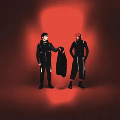Two people in black outfits stand against a red background.