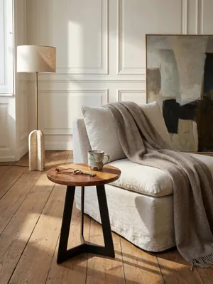 Massiver Holz-Beistelltisch im Industrie-Design arrangiert neben einem tiefen Lounge-Sessel mit drapiertem Kaschmirplaid vor einer warmweißen Wand._#sku_SAFWL5.665#