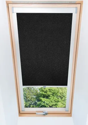 A black roller blind is installed in a wooden-framed attic window, with green trees visible outside.