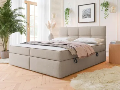 A bedroom scene featuring a boxspring bed with a headboard, mattress, and topper. The bed is dressed with pillows and a throw blanket. A large potted plant is visible to the left, and a small round side table with books is to the right. The room has large
