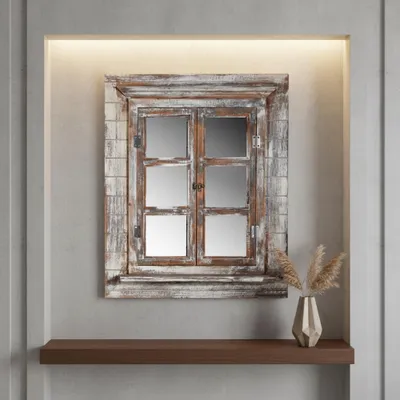 A decorative mirror designed to look like a window with shutters, placed on a wall above a shelf with a vase of dried flowers.