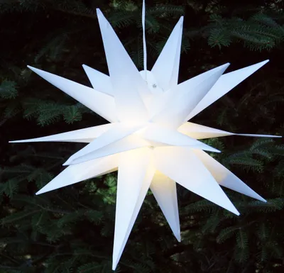 A white, illuminated star-shaped decoration hangs in front of a dark green, out-of-focus background of pine branches.