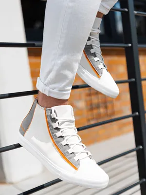 A person wearing white jeans and white high-top sneakers with gray and orange accents is sitting on a black railing.