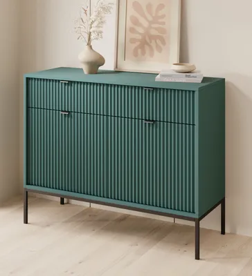 A green dresser with fluted doors and black handles sits on black legs, with a vase and artwork on top.