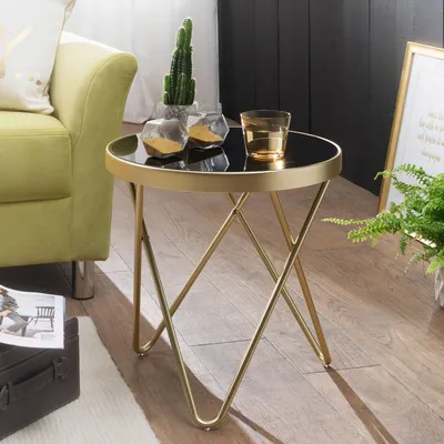 A modern side table with a black top and gold metal legs is shown in a living room setting, next to a green sofa and a potted plant.
