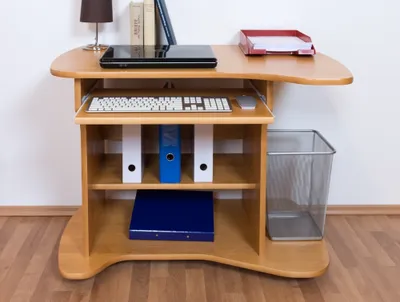 A wooden desk with a laptop, keyboard, mouse, binders, and a trash can.
