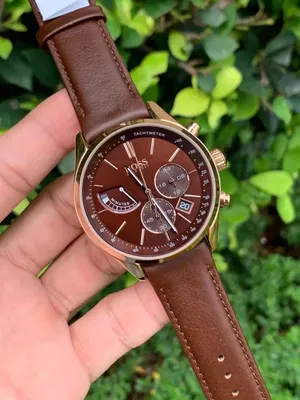 A hand holding a watch with a brown dial and strap, with a green leafy background.