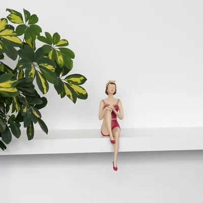 A decorative figurine of a woman in a red swimsuit sitting on a shelf next to a plant.