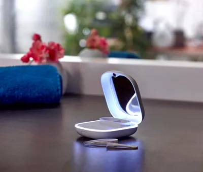 A contact lens cleaner case with a mirror and light is open on a dark surface, with a blue towel and flowers in the background.