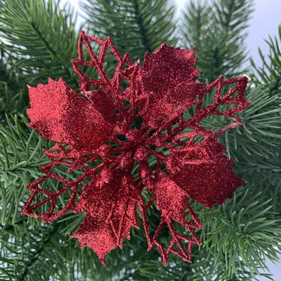 A red glitter poinsettia ornament is placed on a green artificial Christmas tree branch.