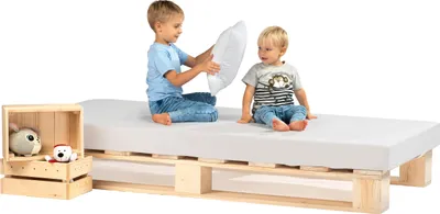 Two young children are playing on a bed frame made of wooden pallets with a mattress on top. A wooden crate with stuffed animals is next to the bed.