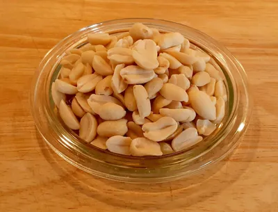 A clear, overhead shot of blanched peanuts in a small glass dish, placed on a wooden surface.