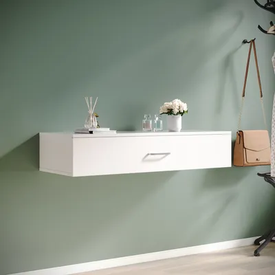 A white floating shelf with a drawer is mounted on a green wall, decorated with a vase of flowers and a handbag hanging on a coat rack.