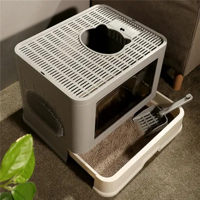 A modern, enclosed cat litter box with a pull-out tray and a litter scoop, placed on a carpeted floor next to a piece of furniture and a plant.
