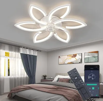 A bedroom with a modern ceiling fan light. The fan has a flower-like design with LED lights. There is a bed in the foreground, and two paintings on the wall. A remote control and a smartphone displaying a control app are in the foreground.