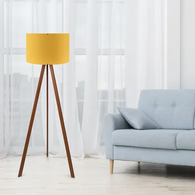 A yellow floor lamp with a wooden tripod base stands next to a light blue sofa in a room with sheer white curtains.