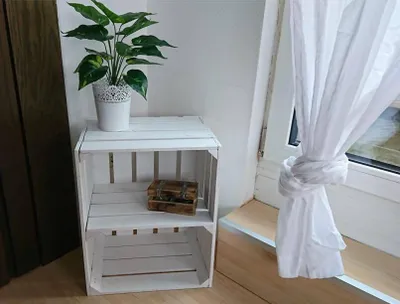 A white wooden crate shelf unit with a plant and a small wooden box on it, placed next to a window with white curtains.