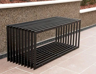 A modern black metal garden bench is placed outdoors on a paved patio, with a textured stone wall and landscaping in the background.