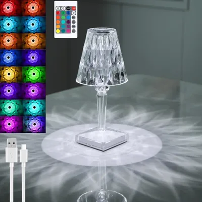 A clear acrylic table lamp with a faceted design, casting light patterns on a reflective surface. A remote control and USB cable are also visible.