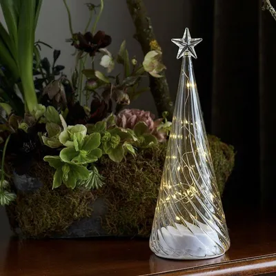 A decorative glass Christmas tree with LED lights sits on a wooden surface next to a planter filled with greenery and flowers.