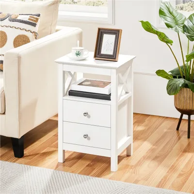 A white nightstand with two drawers is placed next to a sofa in a living room setting. A plant in a gold pot is also visible.