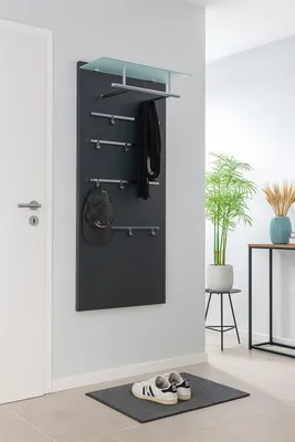 A modern entryway with a black coat rack panel mounted on a light gray wall, featuring hooks for hats and scarves, a glass shelf, and a pair of sneakers on a doormat.