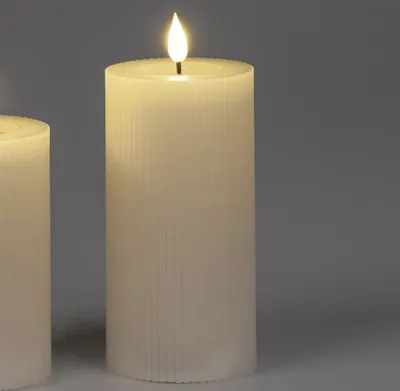 Two cream-colored LED candles with ribbed texture are shown against a plain gray background. The candle in the foreground has a lit LED flame.