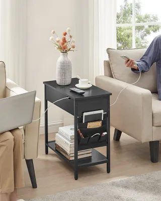 A black side table with storage pockets and charging ports is placed between two chairs in a living room. A person is using a laptop on the left, and another person is using a phone on the right, with charging cables connected to the table.