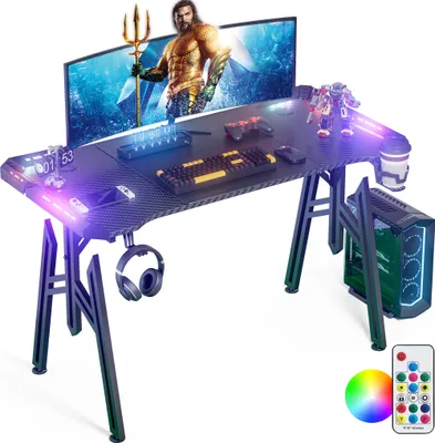 A gaming desk setup with a monitor displaying an image of Aquaman, a keyboard, mouse, game controllers, headphones, a computer tower, and RGB lighting.
