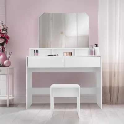 A white vanity table with a tri-fold mirror and a matching stool, set against a pink wall with sheer curtains in the background.