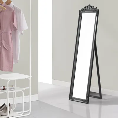 A full-length mirror with a dark gray frame stands on a light gray floor in a room with light gray walls. To the left, a white shelf unit holds shoes and a book, and a clothing rack displays a pink dress and a white shirt.