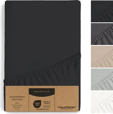 A fitted sheet is shown in black, with a cardboard tag attached. Several other fitted sheets in different colors are displayed vertically on the right side of the image.
