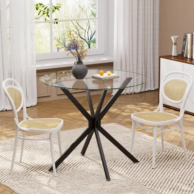 A round glass-top dining table with a black metal base and two white chairs with woven seats are set up in a dining area with a window and curtains in the background.