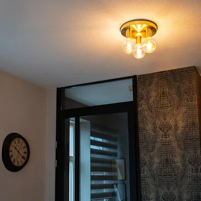 A ceiling light fixture with three bulbs is shown mounted on a white ceiling. To the left, a black wall clock with Roman numerals is visible. To the right, a wall with patterned wallpaper is seen. A glass door with blinds is also in the background.