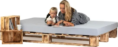 A woman and a child are lying on a bed frame made of wooden pallets, with a mattress on top. There are wooden crates next to the bed.
