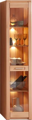 A tall, narrow glass display cabinet made of light-colored wood, with several shelves showcasing decorative items and glassware. The cabinet is illuminated from within.