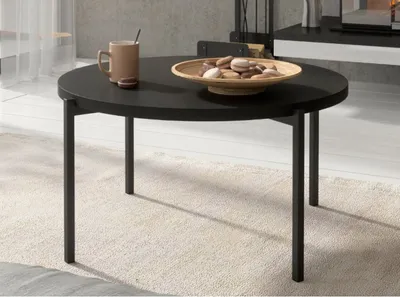 A round black coffee table with a bowl of cookies and a mug on it, placed on a beige rug in a living room setting.