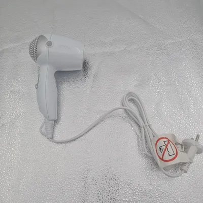 A white Bomann travel hair dryer with a white cord and plug is laid out on a textured, reflective surface.