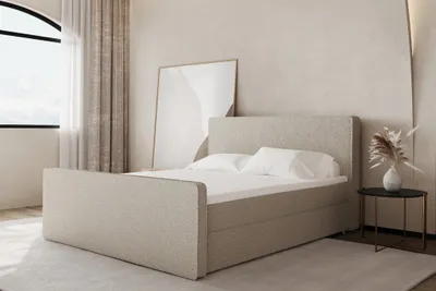A bedroom scene with a beige boxspring bed, a large framed artwork leaning against the wall, and a small side table with a vase of dried flowers.