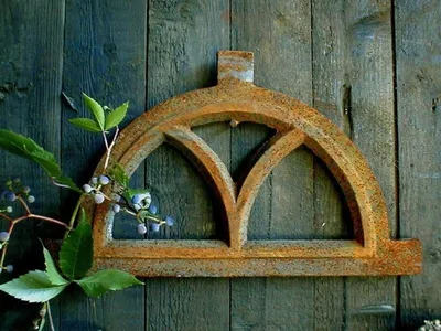 A rusty, arched metal window frame is mounted on a weathered wooden wall, with a branch of green leaves and blue berries in the foreground.