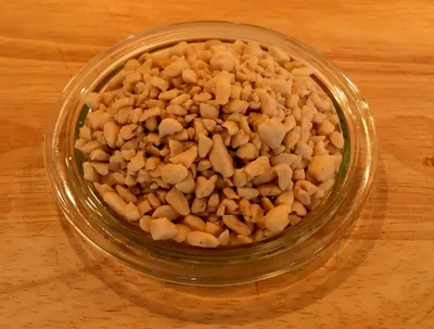 A close-up overhead view of chopped peanuts in a clear glass dish, placed on a wooden surface.
