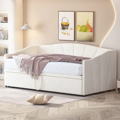 A cream-colored daybed with a pull-out bed is shown in a well-lit room with artwork on the wall and a lamp.